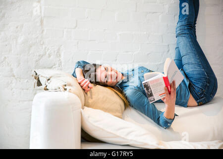 Giovane donna sdraiata sul divano con le gambe sollevate la lettura di un libro Foto Stock