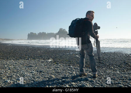 Fotografo maschio sulla spiaggia che porta la telecamera e treppiede, Rialto Beach, nello Stato di Washington, USA Foto Stock
