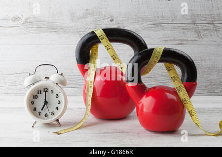 Due kettlebells rosso con nastro di misurazione e vintage orologio sul bianco rustico tavolo in legno. Salute e fitness concetto. Foto Stock