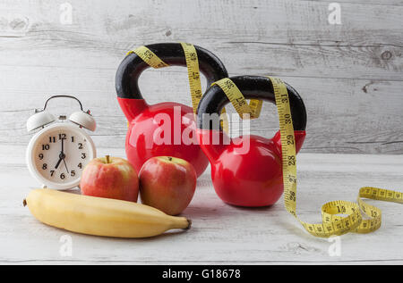 Due kettlebells rosso con nastro di misurazione, mele, banane e vintage orologio sul bianco rustico tavolo in legno. Una dieta salutare e fitnes Foto Stock