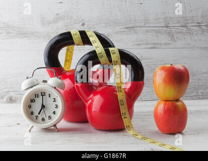 Due kettlebells rosso con nastro di misurazione, mele e vintage orologio sul bianco rustico tavolo in legno. Dieta sana e concep fitness Foto Stock