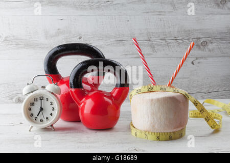 Due kettlebells rosso con nastro di misurazione, bere noce di cocco, cannucce e vintage orologio sul bianco rustico tavolo in legno. Alimentazione sana Foto Stock