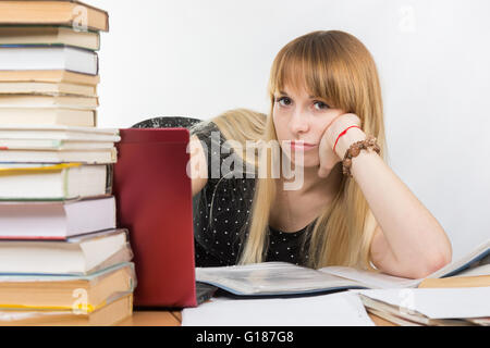 Una studentessa seduti ad una scrivania con un computer portatile e tristemente guarda fuori da dietro una pila di libri Foto Stock