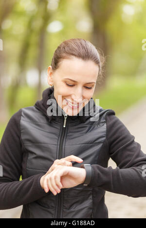 Giovane donna utilizzando smart guarda dopo il jogging per frequenza cardiaca di controllo. Ragazza sorridente con tracker di fitness con la fascetta da polso a all'aperto Foto Stock