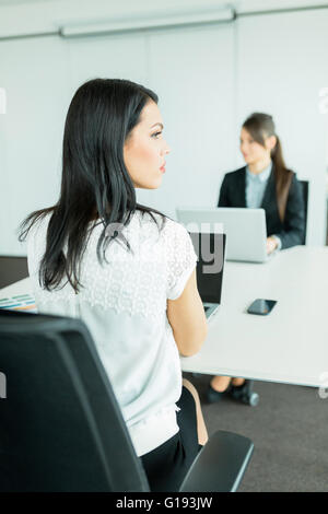 Bella giovane imprenditrice guardando lontano dalla scrivania in un ufficio mentre il suo collega funziona su un computer portatile Foto Stock