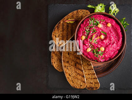 Arrosto di barbabietole hummus con pane tostato in un vaso di ceramica su uno sfondo scuro. Vista superiore Foto Stock