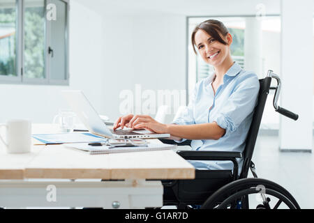Fiducioso felice imprenditrice in carrozzella a lavorare alla scrivania in ufficio e utilizzando un computer portatile, lei è sorridente in telecamera, disabilità oltre Foto Stock