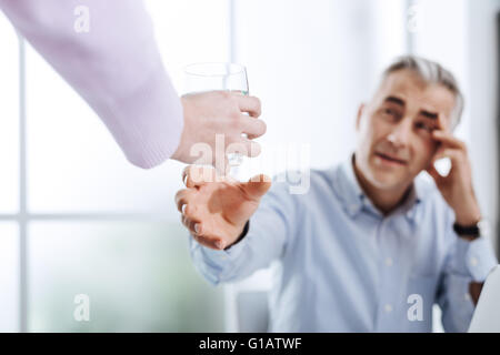 Esaurito imprenditore lavorando nel suo ufficio e con un mal di testa, il suo assistente è di consegnare a lui un bicchiere di acqua, stress e postare Foto Stock
