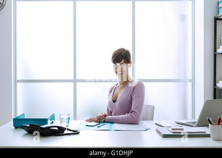 Fiducioso sorridente giovane imprenditrice nel suo ufficio, guarda la fotocamera e seduti alla scrivania Foto Stock