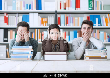 Cieco e sordo e muto gli studenti che copre gli occhi, le orecchie e la bocca, essi sono seduti alla scrivania e poggiando su pile di libri Foto Stock