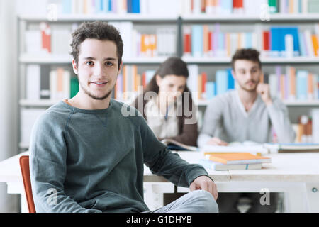 Giovani studenti di sesso maschile presso la biblioteca di sorridere alla telecamera, i suoi compagni di scuola sono seduti alla scrivania sullo sfondo Foto Stock