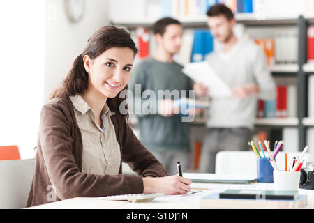 Giovane studentessa di fare i compiti e studiare in biblioteca, gli studenti e gli scaffali su sfondo, l educazione e l apprendimento Foto Stock