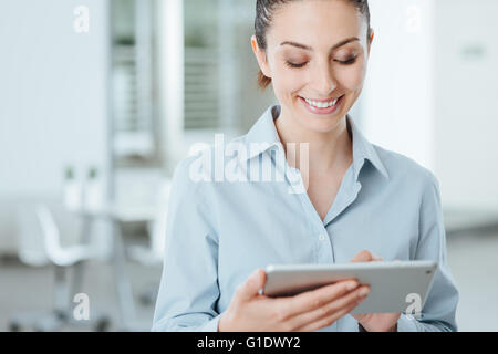 Sorridente giovane donna di affari con un digital tablet touch-screen e utilizzando le app, sta in piedi in ufficio Foto Stock