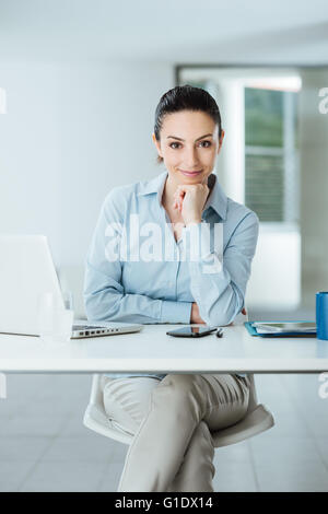 Bella fiducioso donna manager seduto alla scrivania in ufficio e sorridente in telecamera con la mano sul mento, sala interna su sfondo Foto Stock