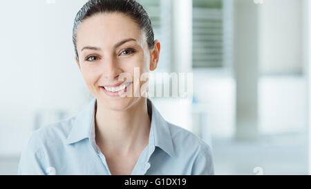 Giovane e bella sorridente business donna sorridente e guardando la telecamera, ufficio interno sullo sfondo Foto Stock