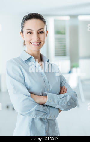 Bella fiducioso business donna posa con bracci incrociati, sorridente in telecamera e in piedi nel suo ufficio Foto Stock