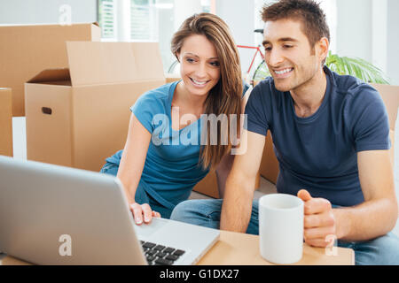 Felice coppia seduta sul loro nuovo piano casa circondata da scatole di cartone e utilizzando un laptop wireless durante una pausa caffè Foto Stock