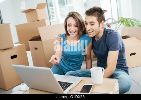 Sorridente giovane seduto sul loro nuovo piano casa circondata da scatole di cartone, stanno guardando un video divertente su un computer portatile e laug Foto Stock