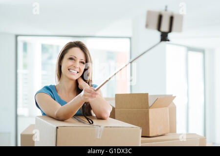 Sorridente giovane donna in movimento nella sua nuova casa e tenendo selfies con un bastone selfie Foto Stock