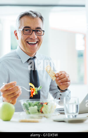 Imprenditore sorridente seduto alla scrivania in ufficio e avente una pausa pranzo, egli è mangiare una insalatiera Foto Stock