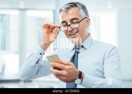 Imprenditore sorridente con problemi di vista, egli sta regolando i suoi occhiali e leggendo qualcosa sul suo telefono cellulare Foto Stock