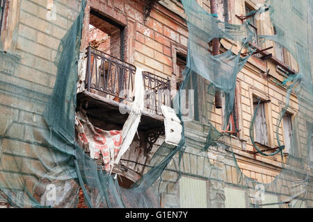 Vecchia casa vivente è in ricostruzione, facciata con balcone rotto è ricoperta di verde maglia di protezione Foto Stock