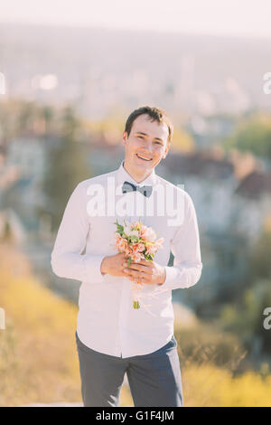 Elegante brunette sorridente lo sposo in camicia bianca con fiocco blu detiene il lussuoso bouquet di fiori all'aperto Foto Stock