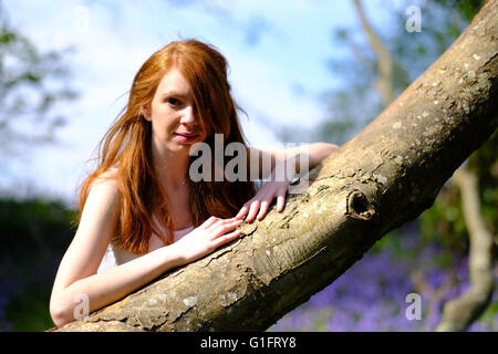 Un esile giovane donna con lunghi capelli rossi si appoggia contro uno spiovente di tronco di albero Foto Stock