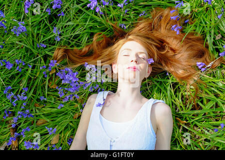 Una snella, equo-pelato i capelli rossi giovane donna che giace tra bluebell fiori a faccia in su. Foto Stock