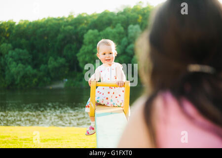 Bambina godendo di equitazione swing con la mamma presso la splendida natura dello sfondo. Baby felicemente giocando con i genitori al di fuori Foto Stock