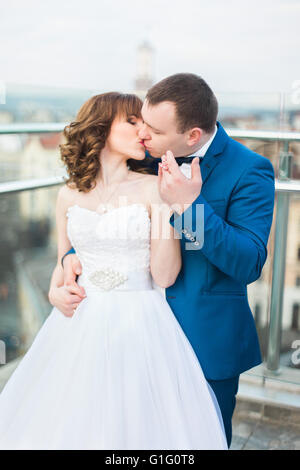 Lo sposo costeggiata dal retro sposa e baciare in piedi sulla terrazza con grande cityscape Foto Stock