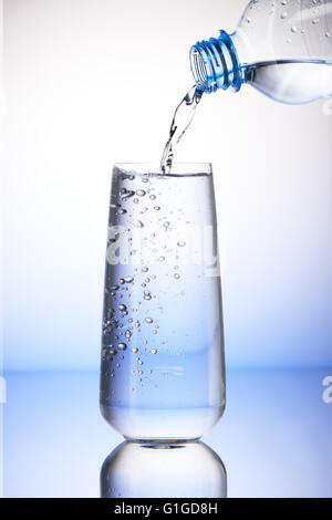 Versare acqua dalla bottiglia di plastica in pieno bicchiere con la riflessione su bianco e lo sfondo blu Foto Stock