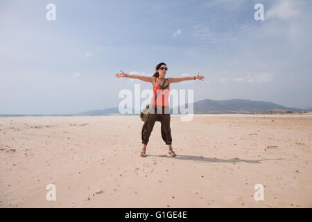 Brunette vacanze estive donna con occhiali da sole, maglietta arancione e marrone e pantaloni in piedi braccia aperte fino abbracciando il saluto di sabbia Foto Stock