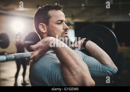 Montare il giovane uomo il sollevamento barbells cercando focalizzata, lavorando in una palestra con altre persone Foto Stock