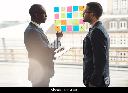 Due imprenditori avente una seria discussione come stanno di fronte a righe di coloratissimi memo su una finestra di vetro per un brainstorming n Foto Stock