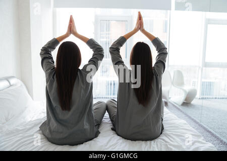 Vista posteriore di due giovani donne in grigio pigiami a praticare yoga e seduta in lotus pongono sul letto Foto Stock