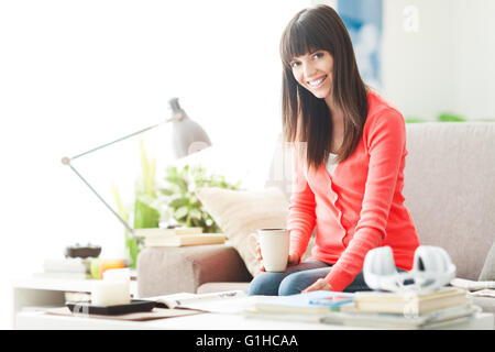 Attraente giovane donna in un momento di relax a casa, lei è seduta sul divano, leggendo una rivista e avente un caffè Foto Stock