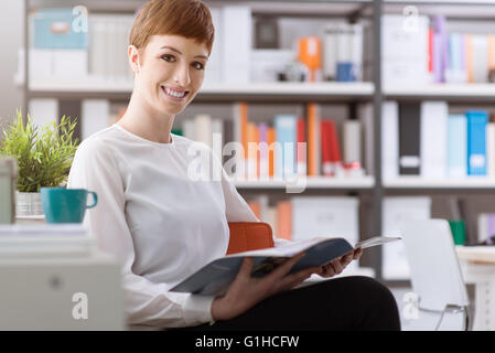 Giovane donna sorridente rilassante, avente un caffè e leggendo una rivista Foto Stock