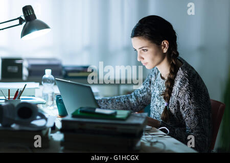 Piuttosto giovane studente seduto alla scrivania e facendo il suo dovere, ella è la connessione a internet con un computer portatile Foto Stock