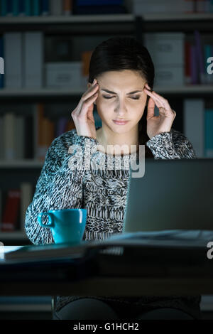 Stanco giovane donna che utilizza un computer portatile e la connessione a internet a tarda notte, sta avendo un mal di testa e toccando il suo templ Foto Stock