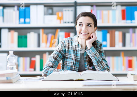 Sorridente ragazza dello studente nella libreria di studiare e di giorno di sognare, ella è pensare con la mano sul mento e guardando in alto, istruzione e Foto Stock