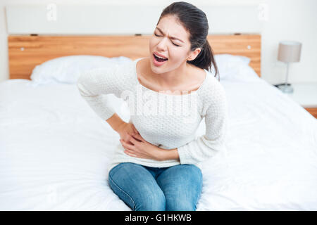 Donna seduta sul letto e toccando il suo lato sinistro nel dolore a casa Foto Stock