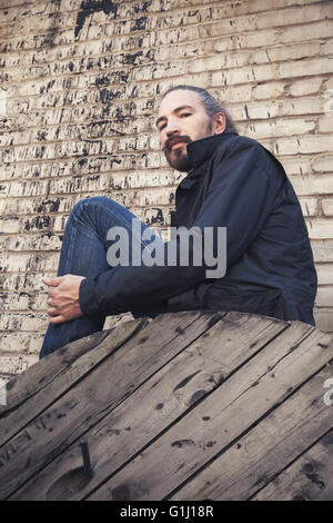 Outdoor Ritratto di giovane barbuto uomo asiatico in nero seduto sul vecchio di legno bobina del cavo Foto Stock