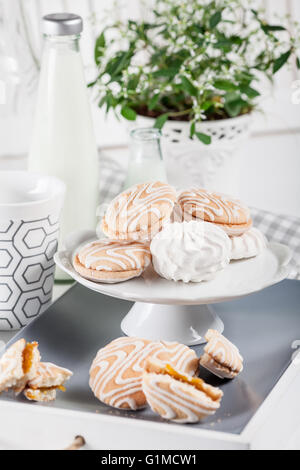 I cookie su bianco e grigio con piastra di tazze di caffè e bottiglie di latte su persiane bianche con sfondo verde e fiore bianco Foto Stock