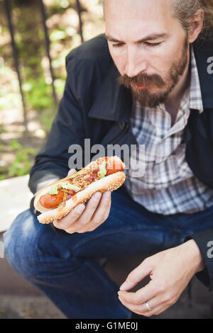 Barbuto uomo asiatico mangiare hot dog in estate park, outdoor ritratto con il fuoco selettivo Foto Stock