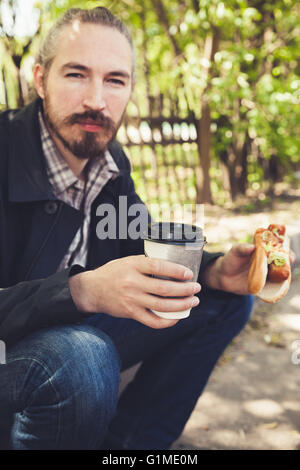 Barbuto uomo asiatico a pranzo con hot dog e caffè nel parco, outdoor ritratto con il fuoco selettivo Foto Stock