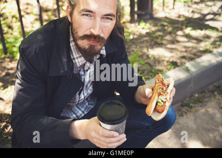 Barbuto uomo asiatico con hot dog e caffè in estate park, outdoor ritratto con il fuoco selettivo Foto Stock