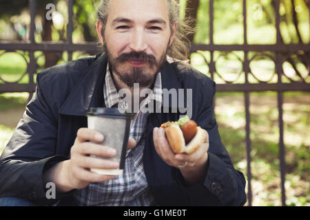 Barbuto uomo asiatico mangiare hot dog con caffè in estate park, outdoor ritratto con il fuoco selettivo Foto Stock