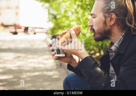 Barbuto uomo asiatico mangiare hot dog con caffè in estate park, il profilo esterno ritratto con il fuoco selettivo Foto Stock