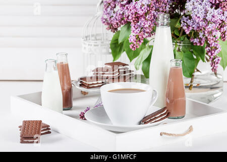 Tazza di caffè e poche bottiglie di latte e cioccolato miklshakes su un vassoio di bianco con fiori lilla bianco su sfondo serrande Foto Stock